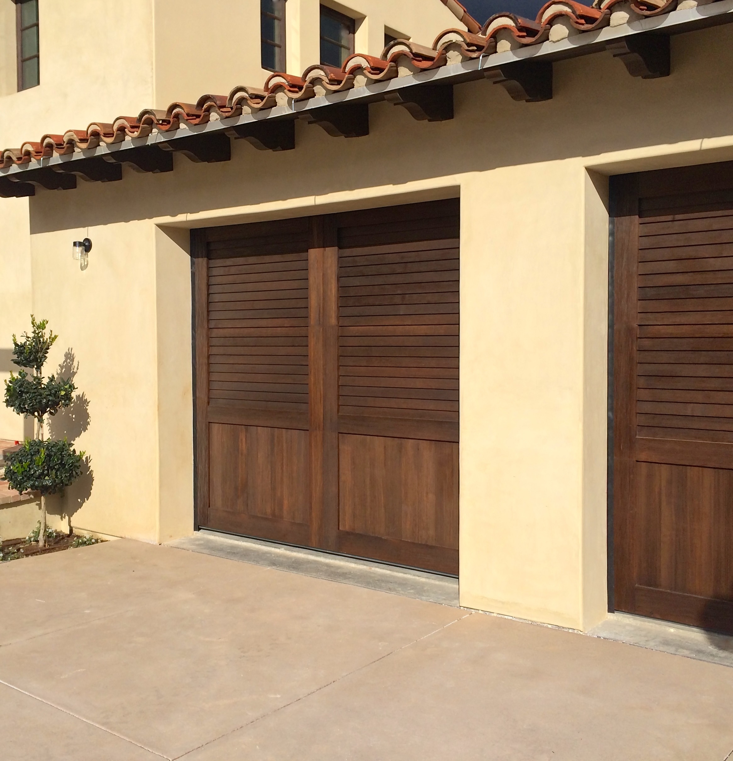 Two closed wooden garage doors with black handles on a stone house exterior, flanked by wall-mounted lanterns and potted plants.