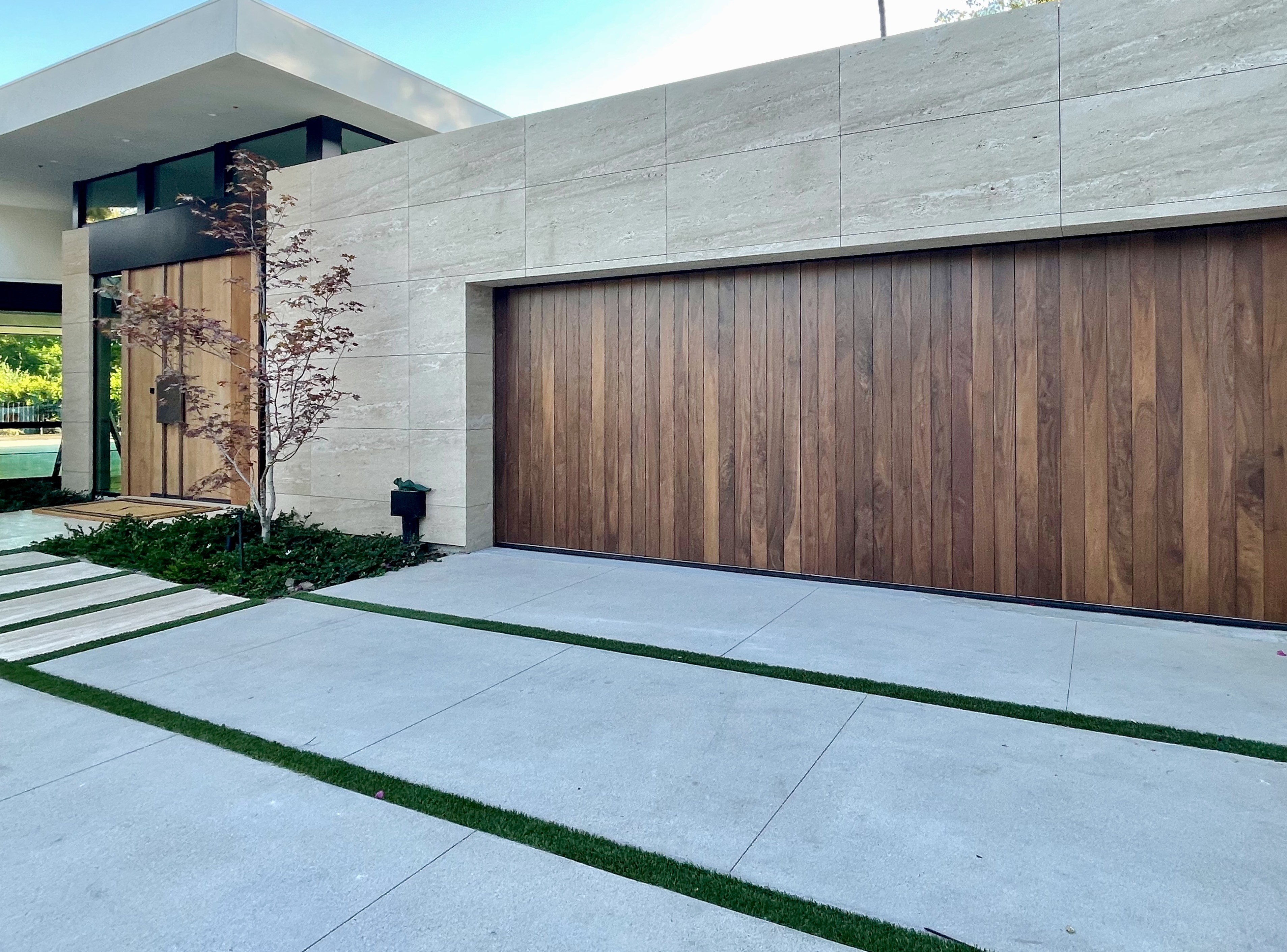Two brown dogs in an open garage with gray double doors and white house exterior, flanked by parked cars.