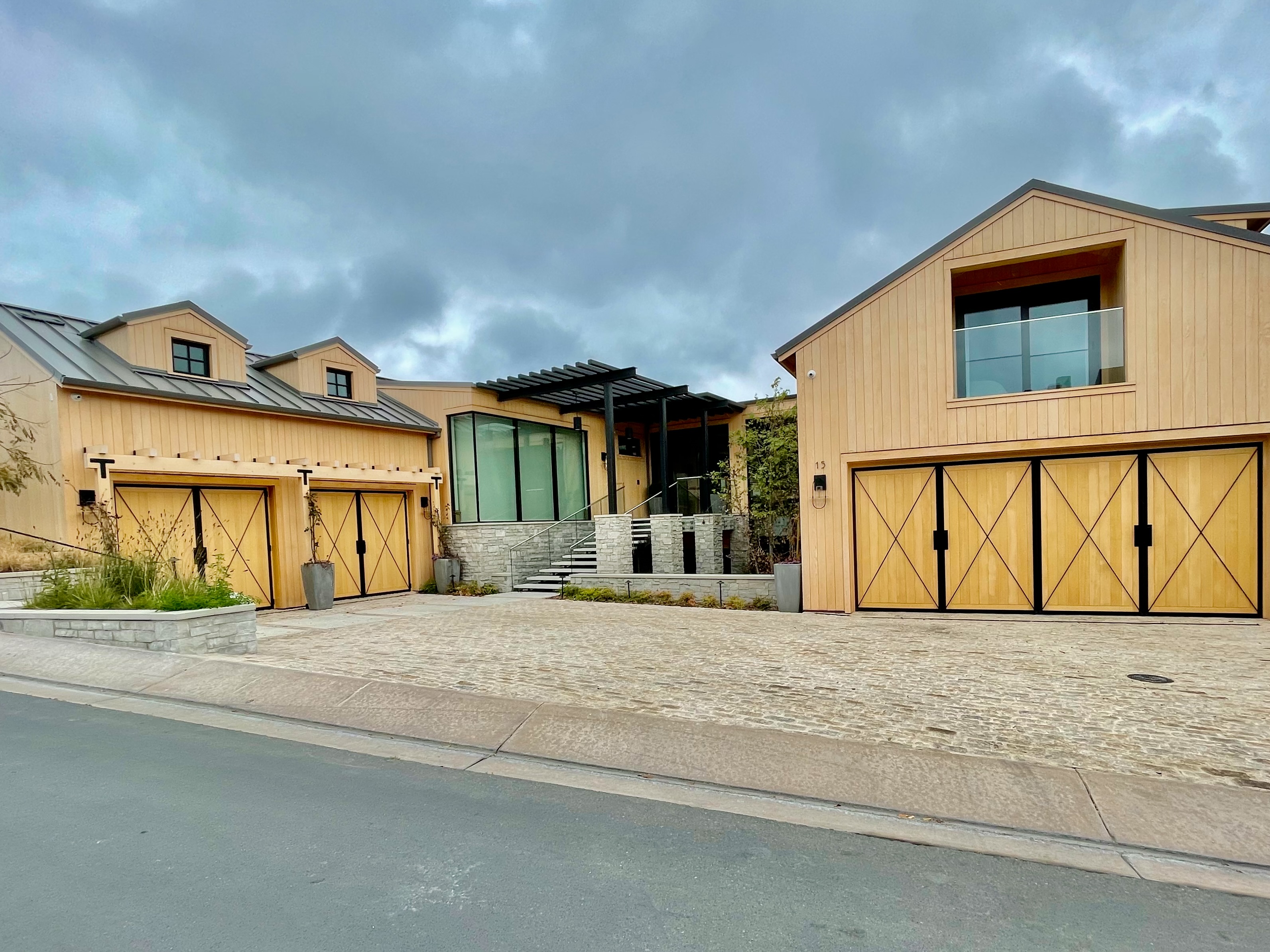 Modern house exterior with a large wooden-framed glass garage door, gray siding, stone chimney, and driveway with a black car partially visible.
