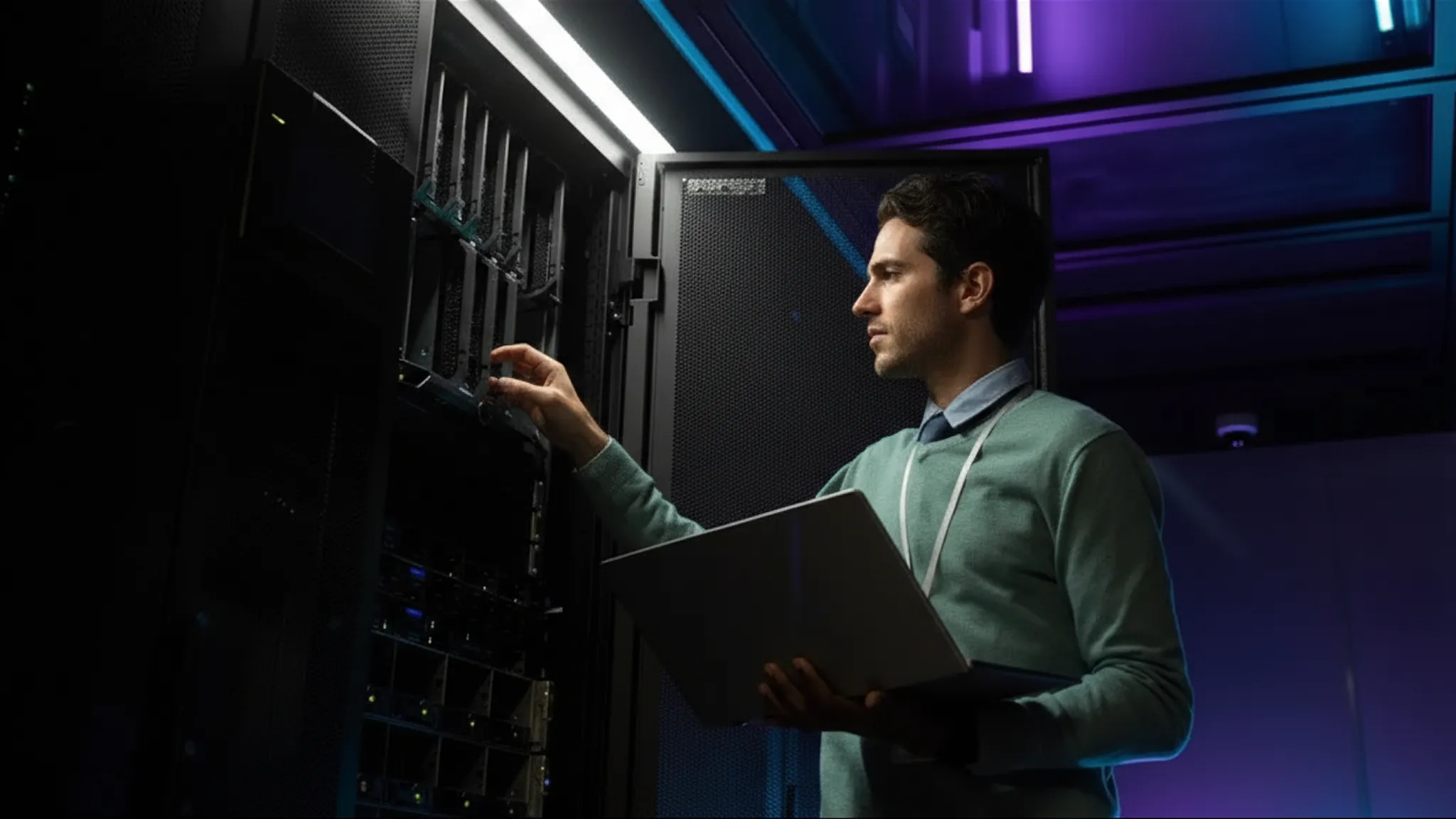 Man holding a laptop and working on a server rack in a dimly lit data center with purple and blue lighting.