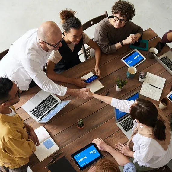 A diverse group of coworkers collaborating around a wooden table with laptops, tablets, notebooks, and coffee cups, two shaking hands.