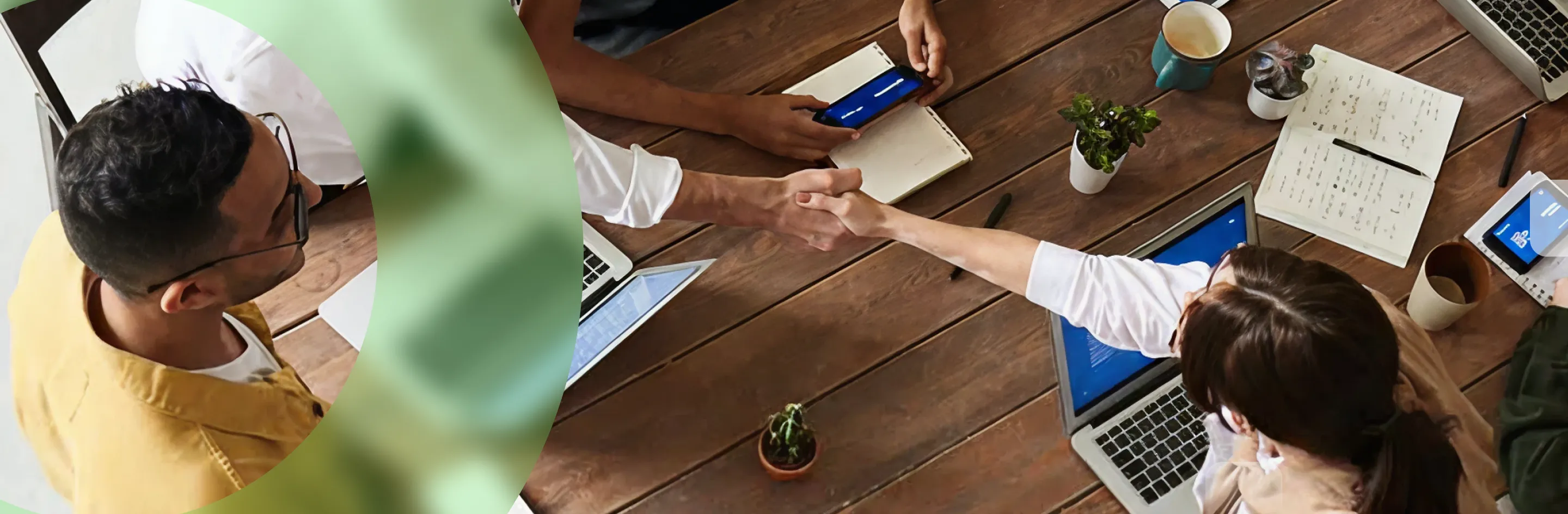Two people shaking hands over a wooden table with laptops, notebooks, plants, and coffee cups.