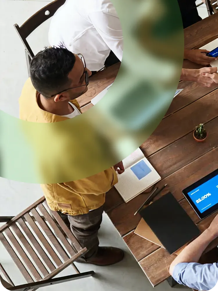 Two people shaking hands over a wooden table with laptops, notebooks, plants, and coffee cups.