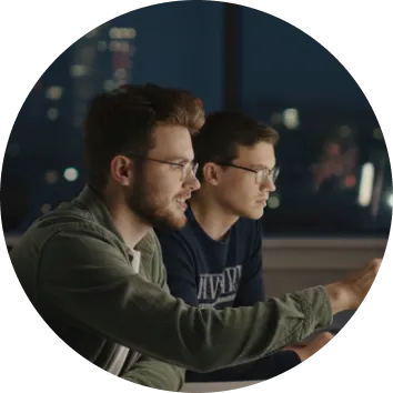 Two men wearing glasses focused on a computer screen in a dimly lit room at night.
