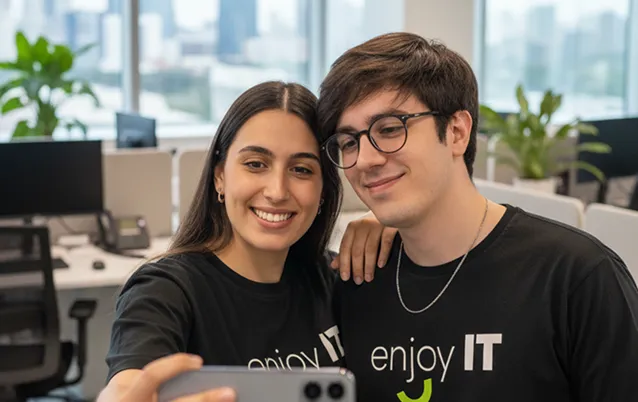 Smiling young man and woman wearing black Enjoy IT t-shirts taking a selfie in an office.