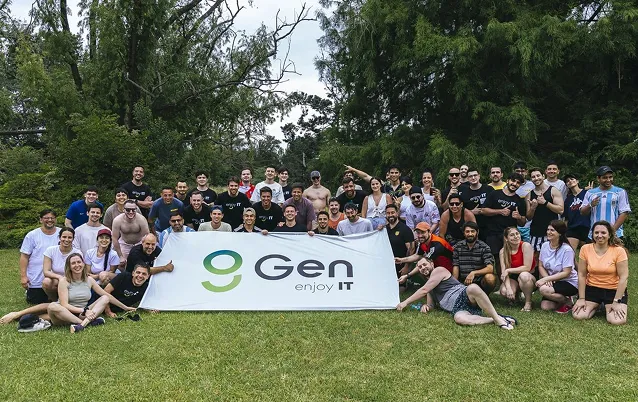 Group of around 40 people posing outdoors on grass behind a banner that reads 'Gen enjoy IT' with trees in the background.