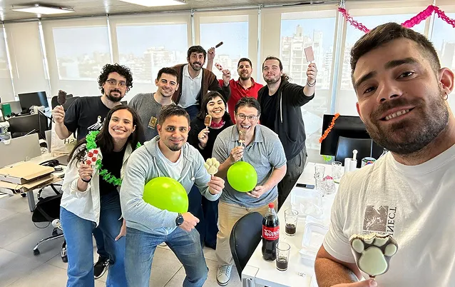 Group of smiling coworkers holding ice cream bars and two green balloons in a brightly lit office setting.