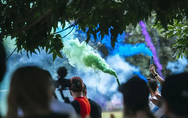 People holding smoke bombs emitting green, blue, and purple smoke outdoors under tree branches.