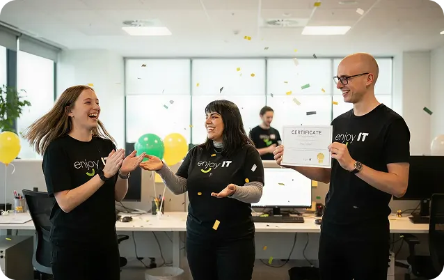 Three people in black enjoy IT shirts celebrating with confetti, one holding a certificate, under a congratulatory banner in an office.