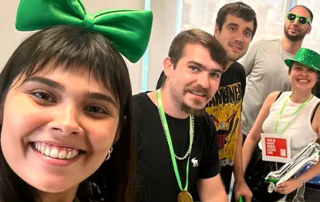 Group of five smiling young people wearing green accessories, celebrating indoors with festive hats and beads.