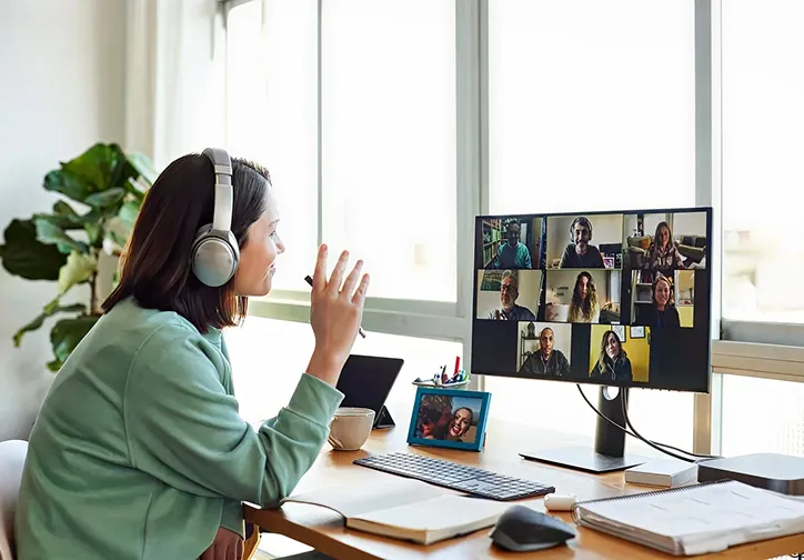 Woman wearing headphones engaging in a virtual meeting on a computer screen with eight participants visible.