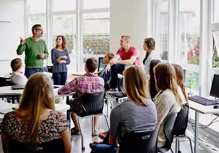 A man and woman standing and speaking to a group of seated young adults in a bright classroom with large windows.