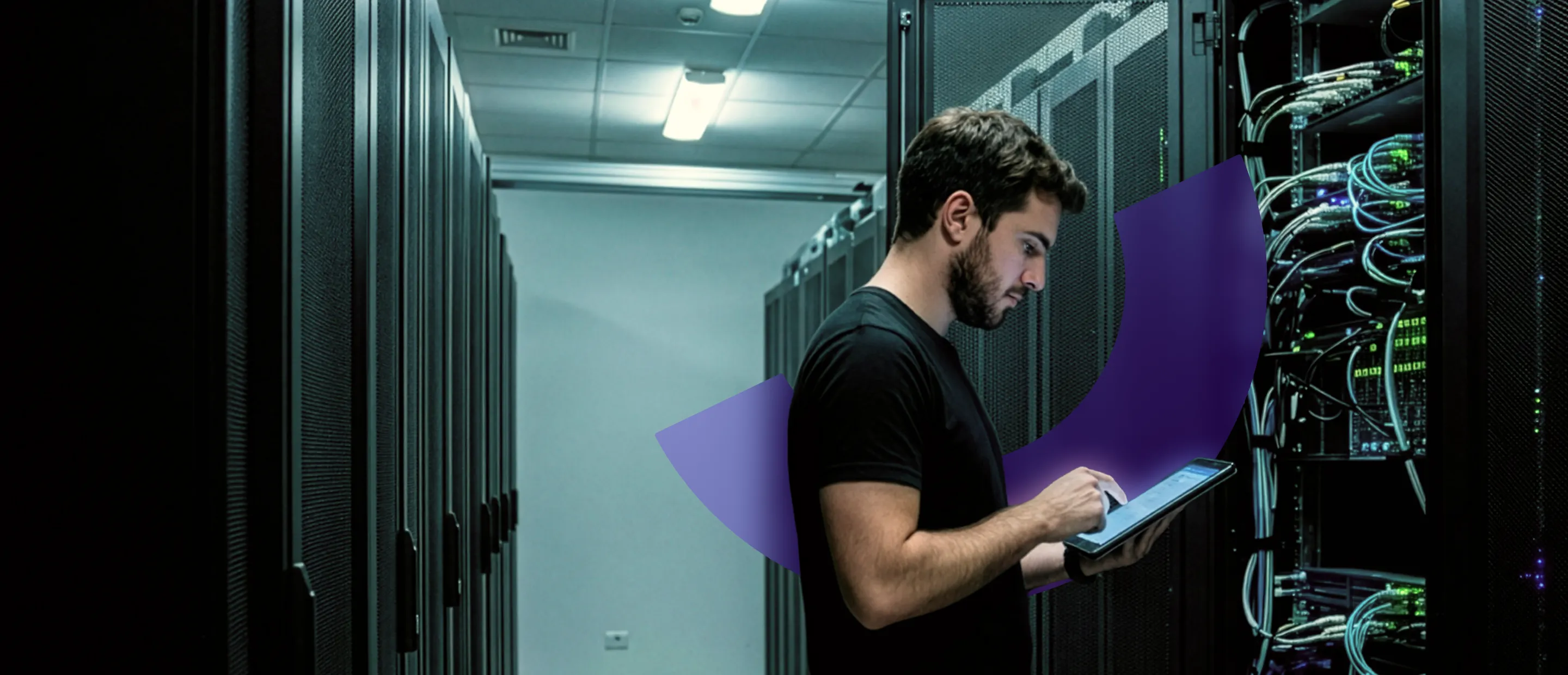 Man in black shirt using a tablet while standing beside server racks in a data center.