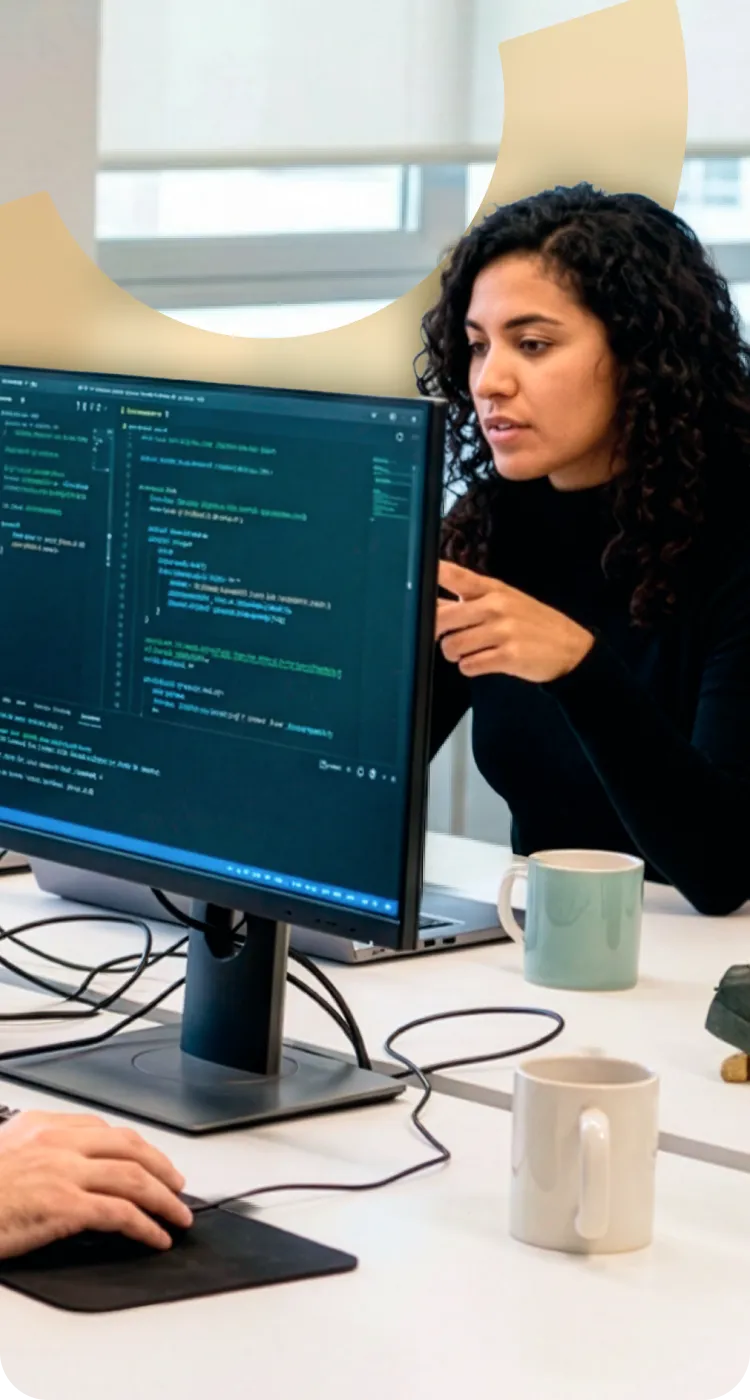 Woman with curly hair pointing at a computer screen displaying code during a discussion with a coworker.