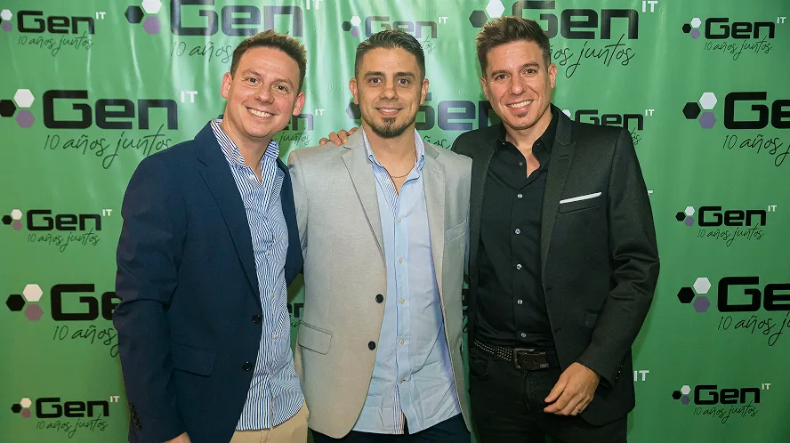 Group of smiling colleagues holding a large white banner with the Gen enjoy IT logo in a bright office.