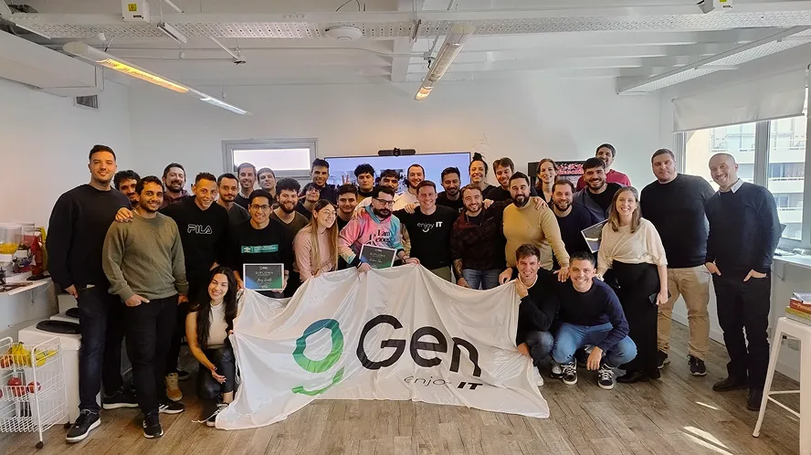 Group of diverse people in casual clothing smiling and holding a white banner with the green and black 'Gen enjoy IT' logo in a modern office setting.