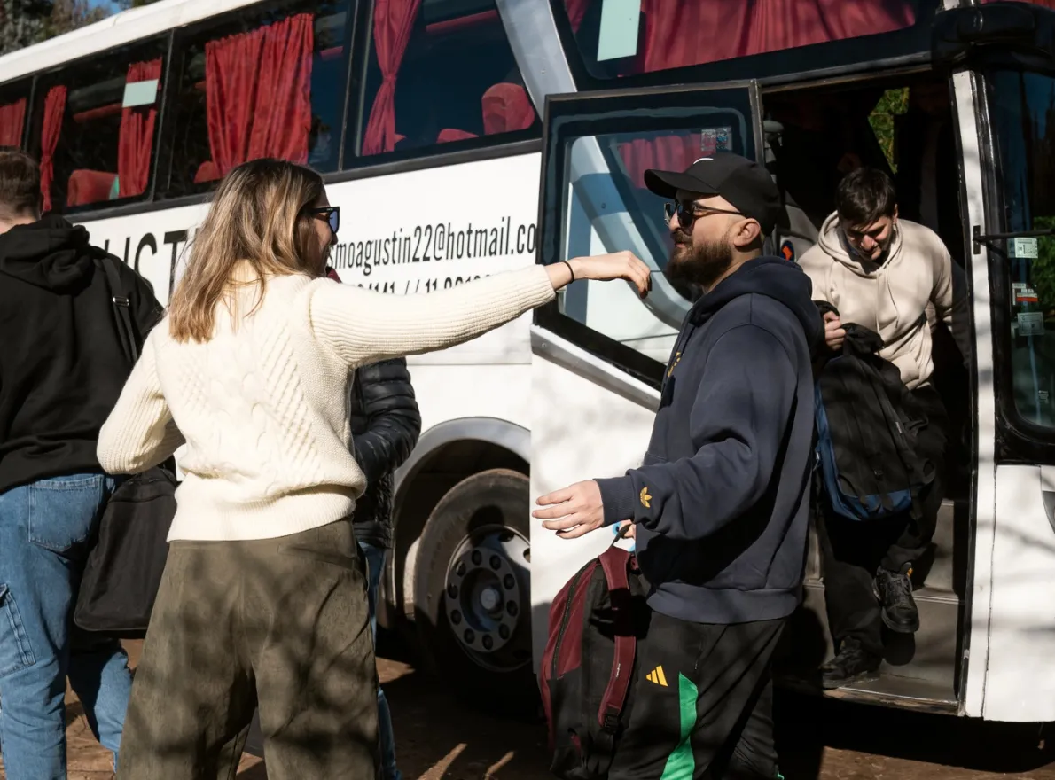 Two people smiling and posing for a selfie outdoors in snowy mountains, one wearing a colorful patterned jacket and green beanie, the other in a grey and blue winter jacket.