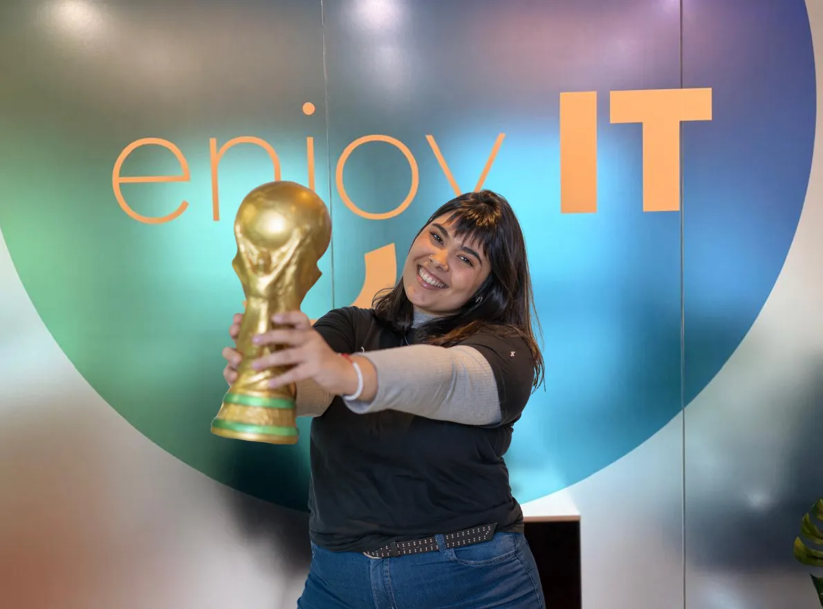 Five young adults smiling and celebrating under a green banner that reads 'enjoy IT' with confetti in the air, all wearing black 'enjoy IT' t-shirts.