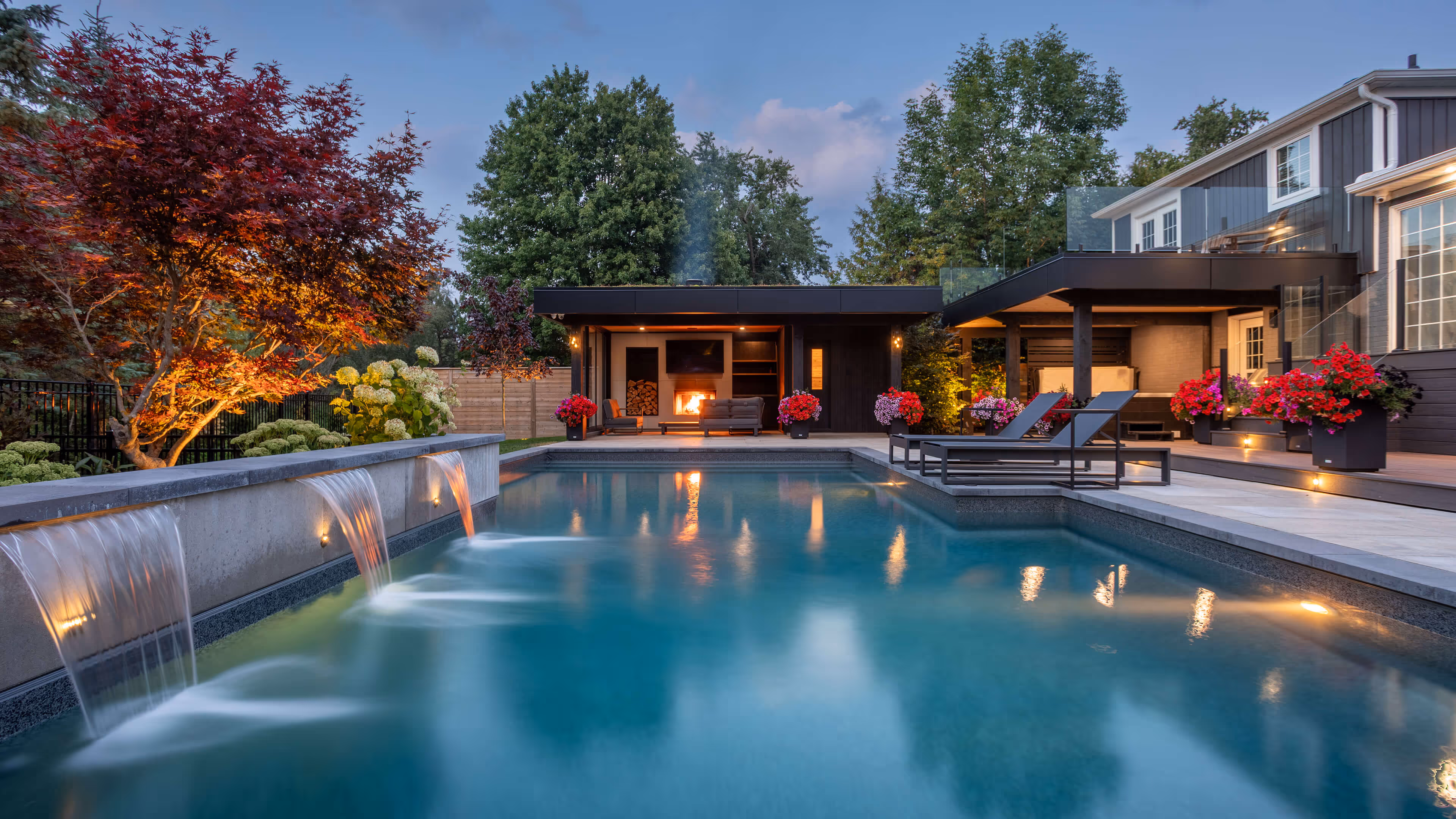 Modern backyard with a swimming pool featuring three waterfalls, lounge chairs, outdoor fireplace, and colorful flower planters.