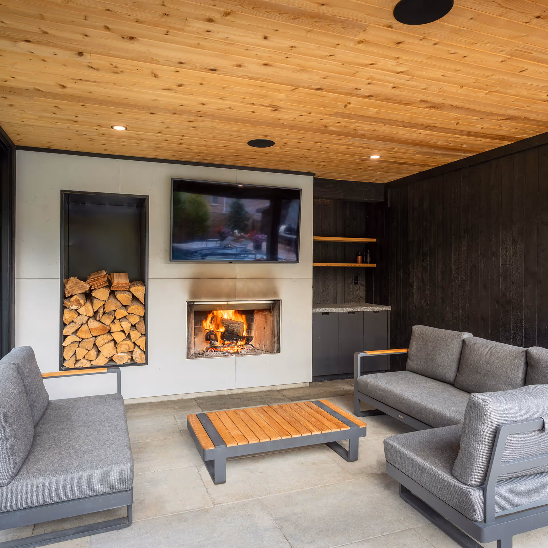 Modern outdoor seating area with grey cushioned sofas, wooden coffee table, fireplace with stacked firewood, and TV mounted above.