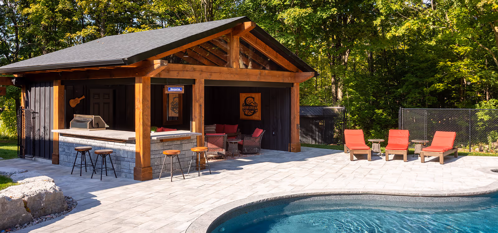 Outdoor poolside pavilion with bar stools, seating area, and three red cushioned lounge chairs on stone patio surrounded by trees.