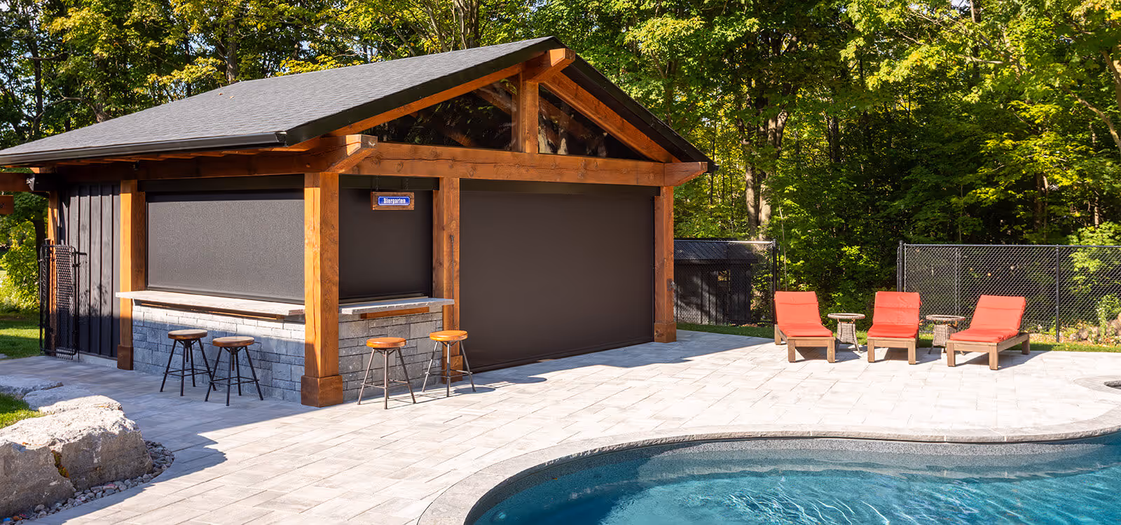 Outdoor pool area with stone patio, three red cushioned lounge chairs, side tables, and a wooden pool house with bar stools.