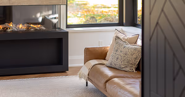 Cozy living room corner with a brown leather sofa adorned with throw pillows and a throw blanket near a modern gas fireplace and large windows.
