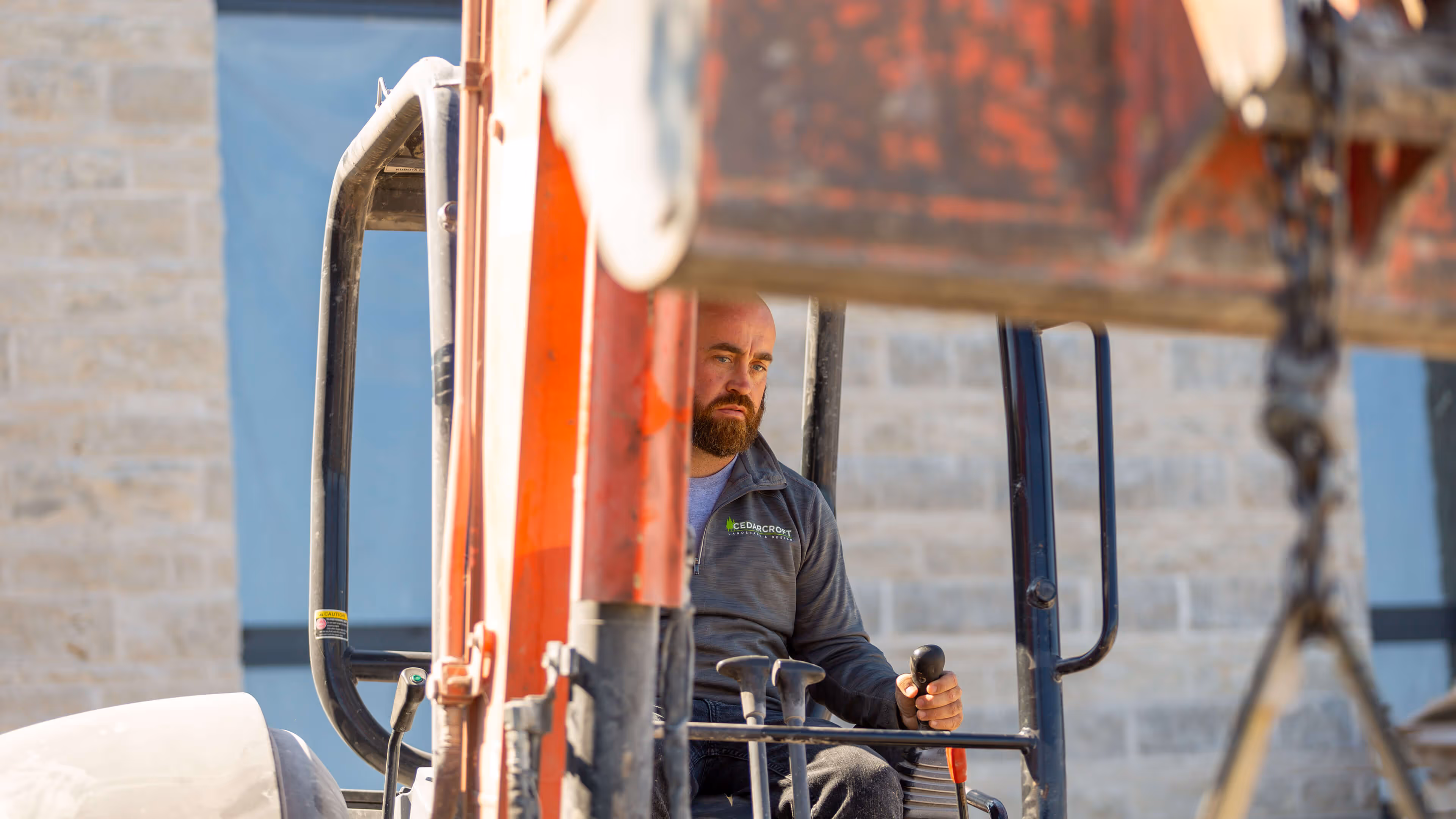 Man with beard operating controls of orange construction equipment outdoors.