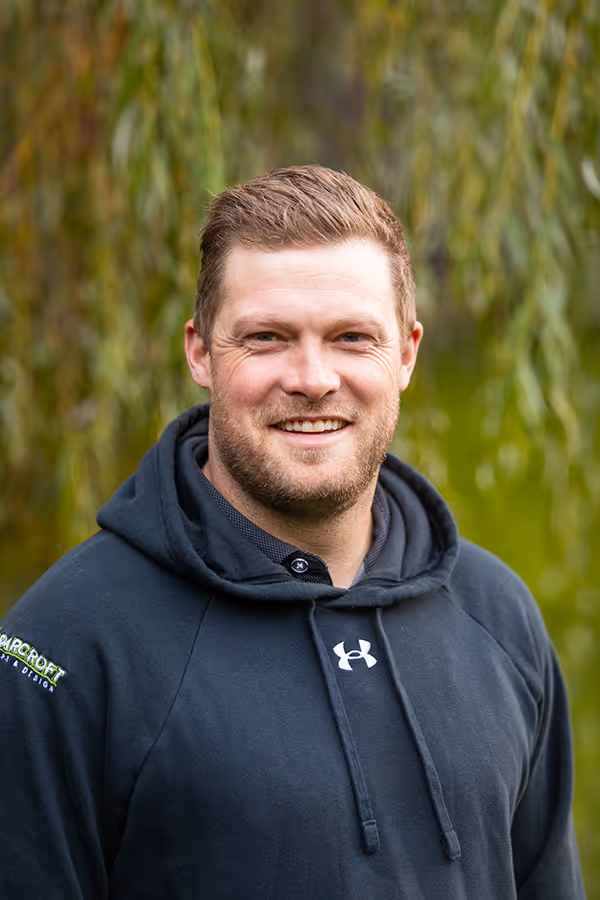 Smiling man with short hair and beard wearing a black Under Armour hoodie outdoors with blurred green foliage background.