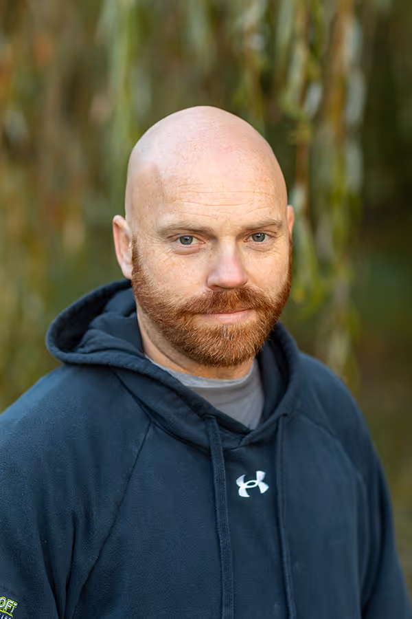 Bald man with a red beard wearing a black Under Armour hoodie standing outdoors with blurred green foliage background.