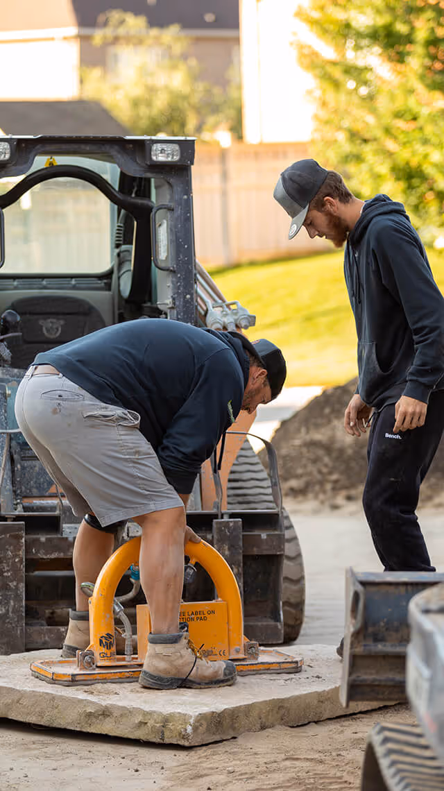 Two construction workers, one bending over operating a yellow tool on a large stone slab, with a small loader vehicle in the background.