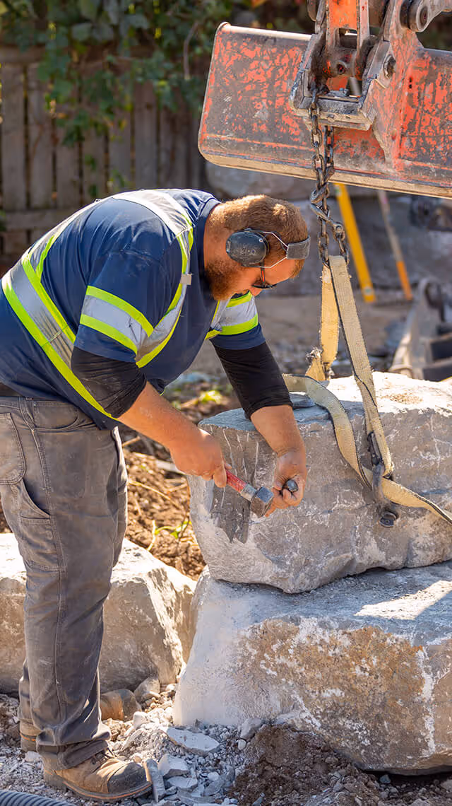 Construction worker wearing ear protection chiseling a large stone block suspended by straps on a construction site.