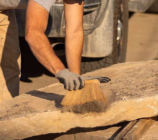 Person wearing a glove brushing dust off a large stone slab outdoors near a vehicle.
