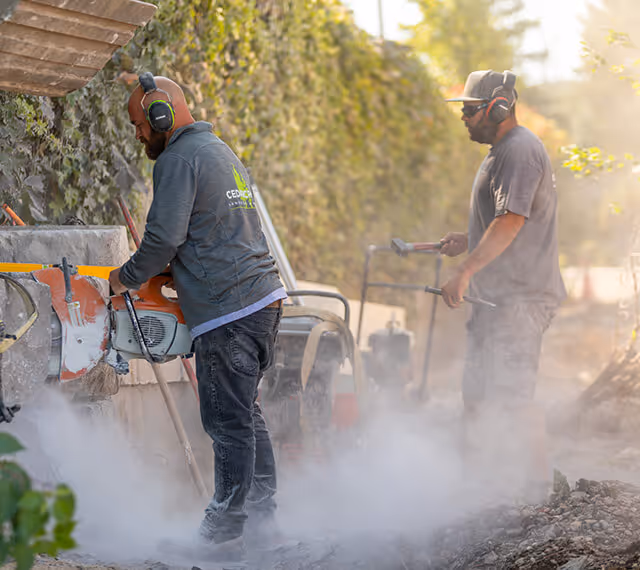 Two construction workers wearing protective headphones operating equipment amid dust and debris outdoors.