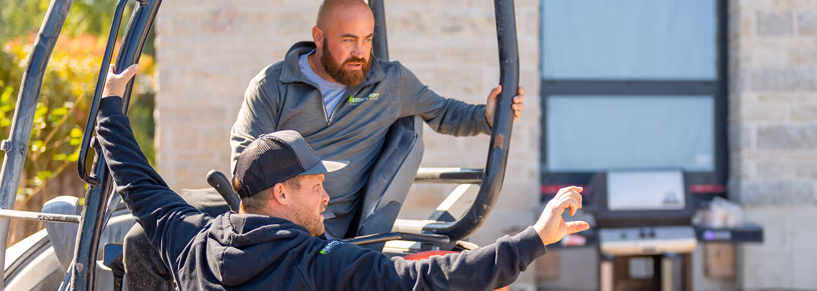 Two men in outdoor workwear on construction equipment, one gesturing with hand while the other listens.