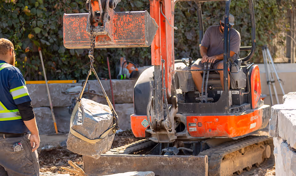 Construction worker standing near an orange mini excavator lifting a large rock with a sling.