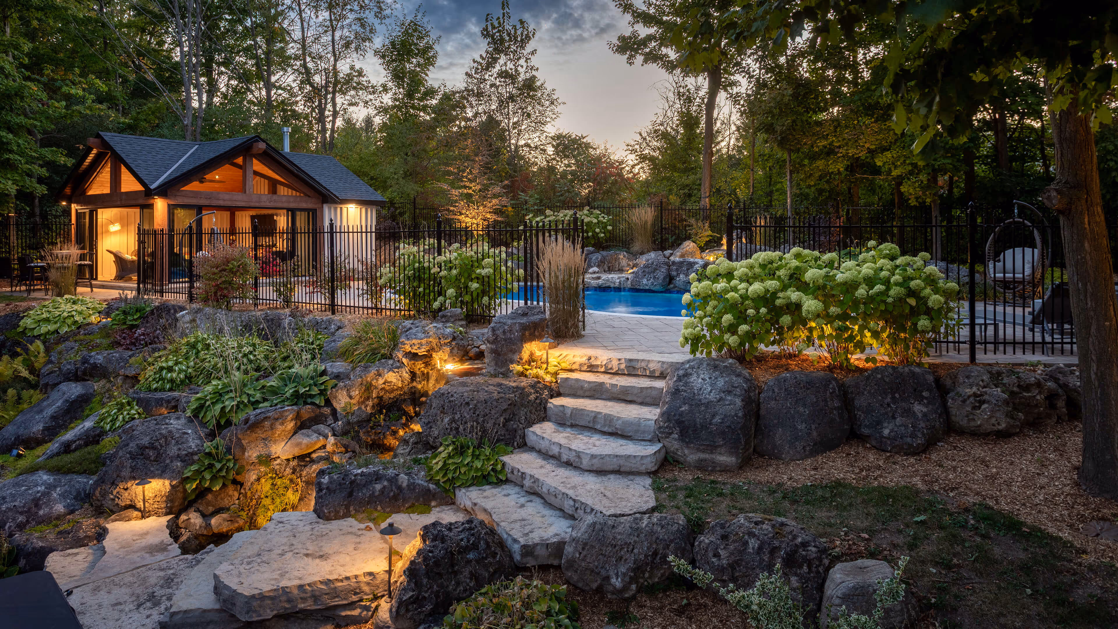 Stone stairs leading to a landscaped backyard with lush greenery, flowers, a fenced pool, and a warmly lit modern pool house at dusk.