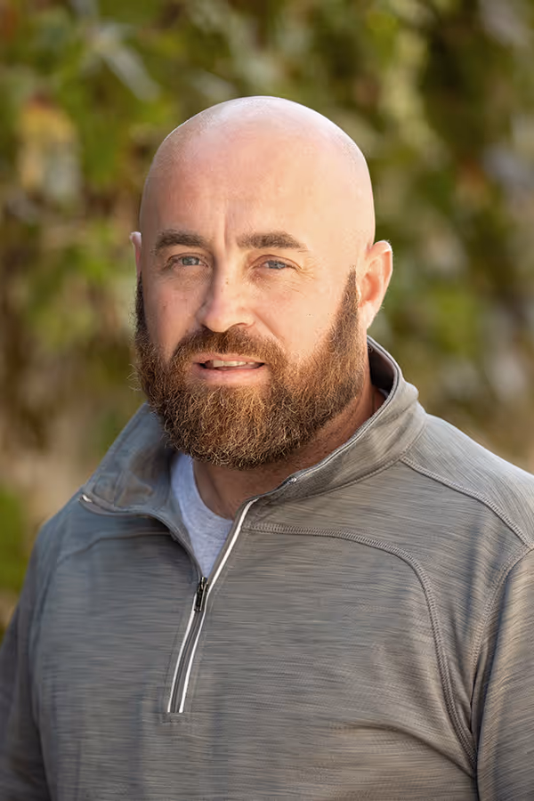 Portrait of a bald man with a full beard wearing a gray quarter-zip pullover, with blurred green foliage background.
