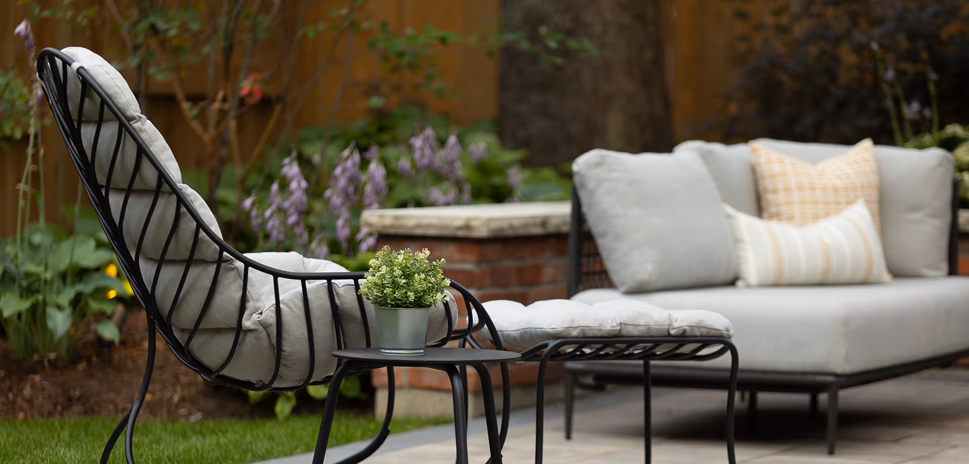 Outdoor patio seating with cushioned black metal chair, matching ottoman, small black side table with a plant, and a cushioned sofa with decorative pillows in a garden setting.