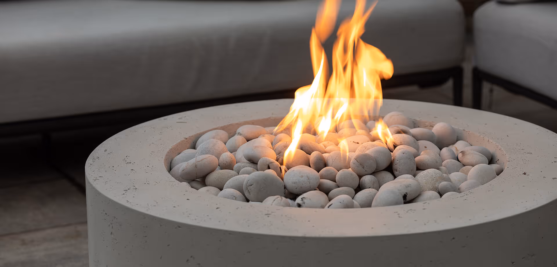 Modern round fire pit with smooth white stones and bright flames, surrounded by gray outdoor seating.