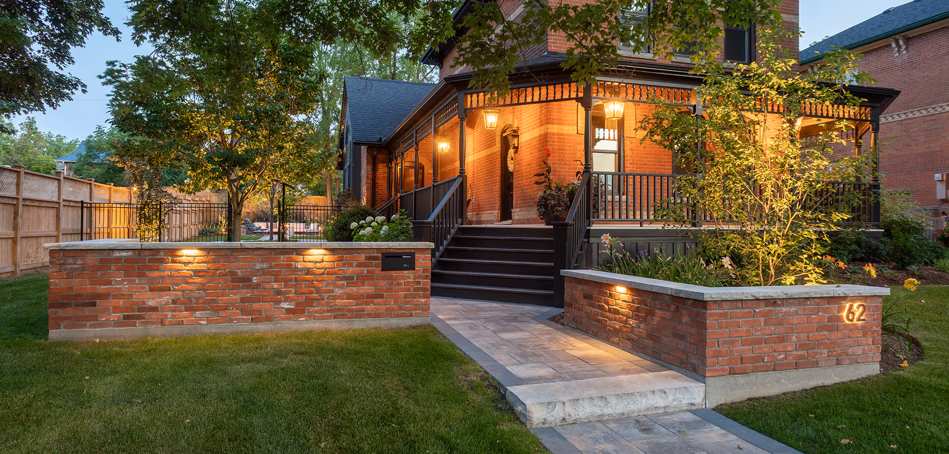 Brick house with lit porch lights, black stairs, and a paved walkway bordered by illuminated brick planters and green lawn.