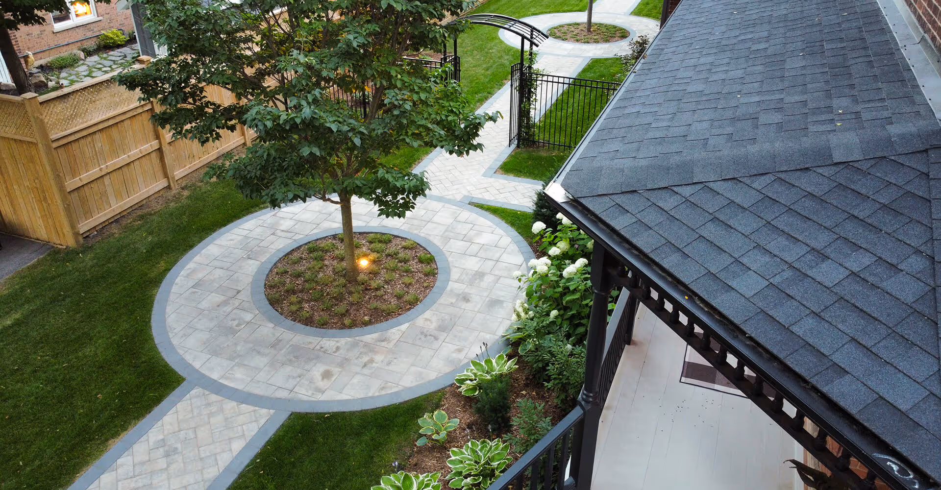 Overhead view of a paved circular patio with a tree in the center, surrounded by a green lawn, garden plants, and a black metal gate with an archway.