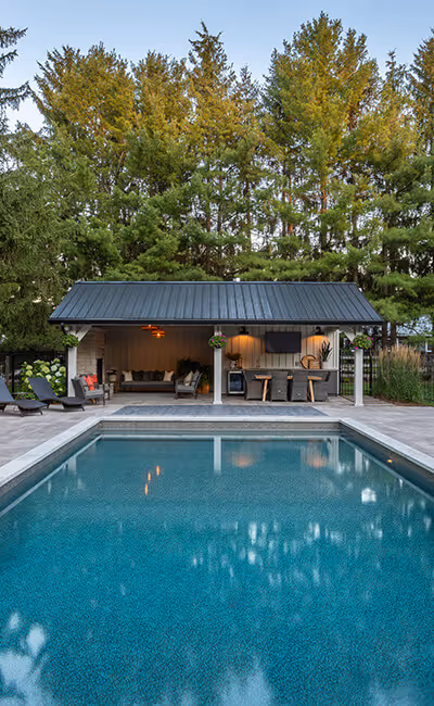 Rectangular outdoor swimming pool with lounge chairs and a covered seating area with bar stools, surrounded by tall trees.