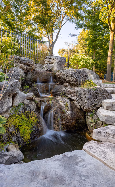 Small multi-tiered waterfall flowing over moss-covered rocks next to stone steps in a leafy garden.