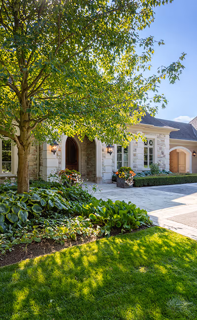 Sunlit stone house with arched doorway, flower pots, lush green lawn, and leafy tree in front garden.