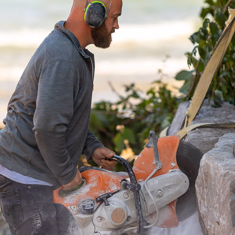 Man wearing ear protection operating a large orange stone-cutting saw near rocky surface outdoors.
