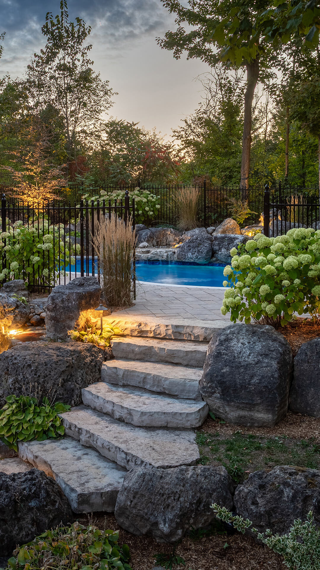 Stone steps leading up to a landscaped backyard with a swimming pool, surrounded by rocks, hydrangeas, and trees at dusk.