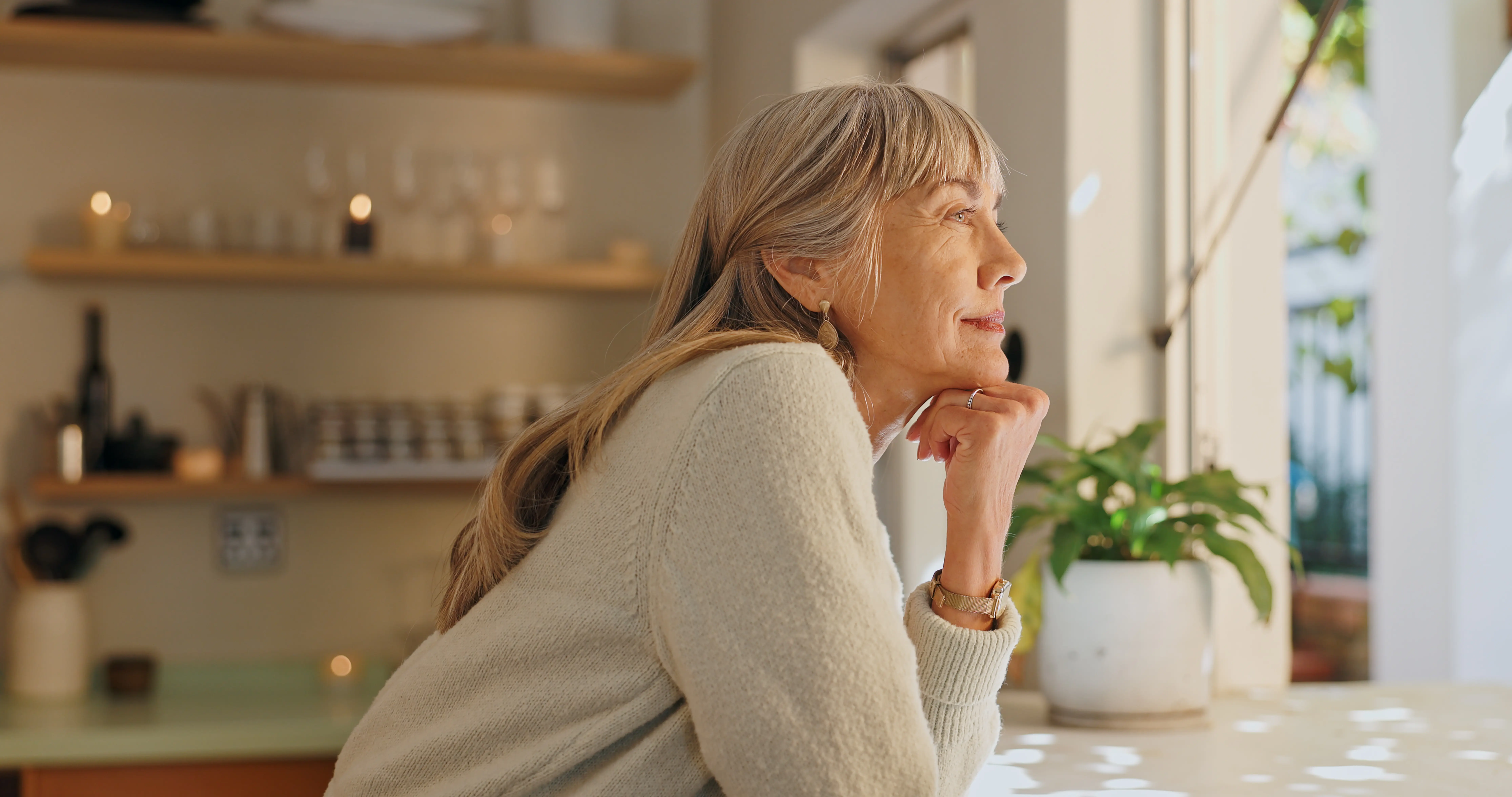 Thoughtful older woman in a light sweater resting her chin on her hand while looking out a sunlit window.