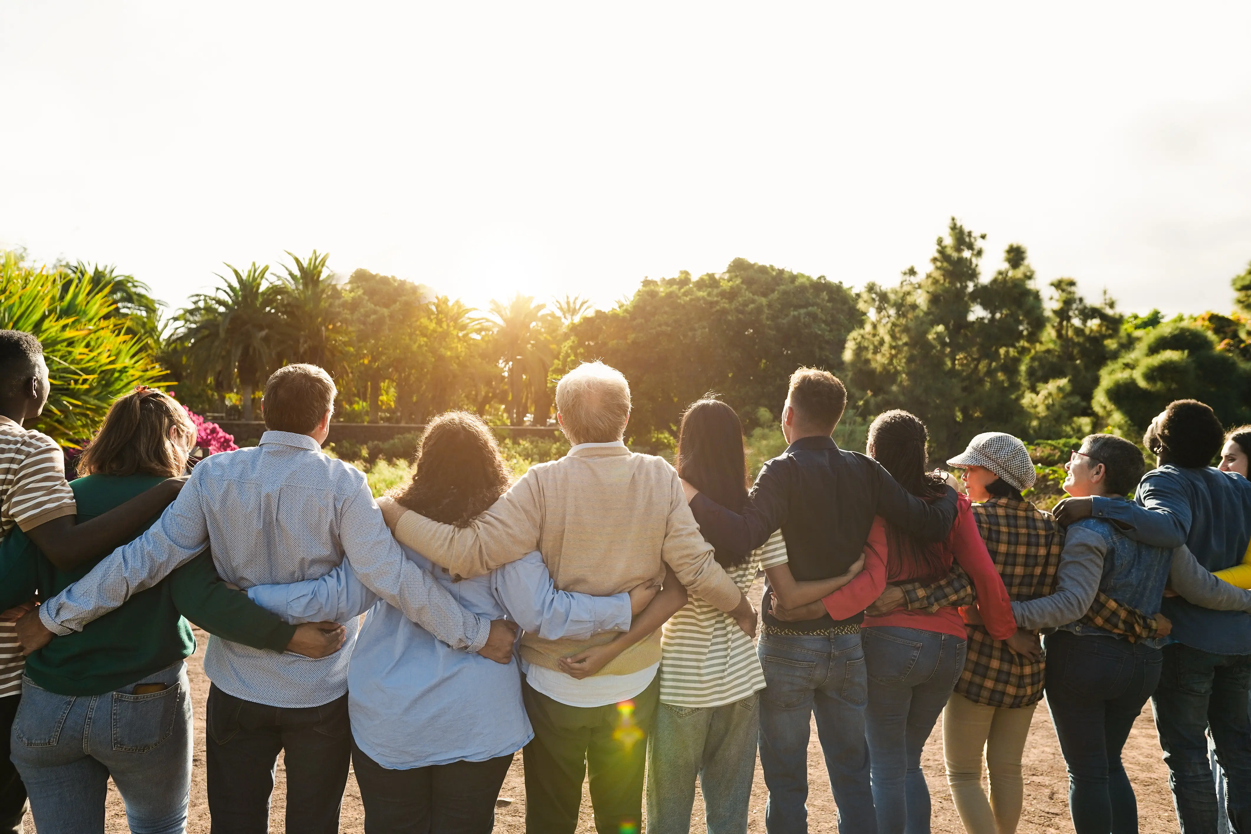 Diverse group of people standing outdoors with arms around each other, facing away toward a sunlit green landscape.