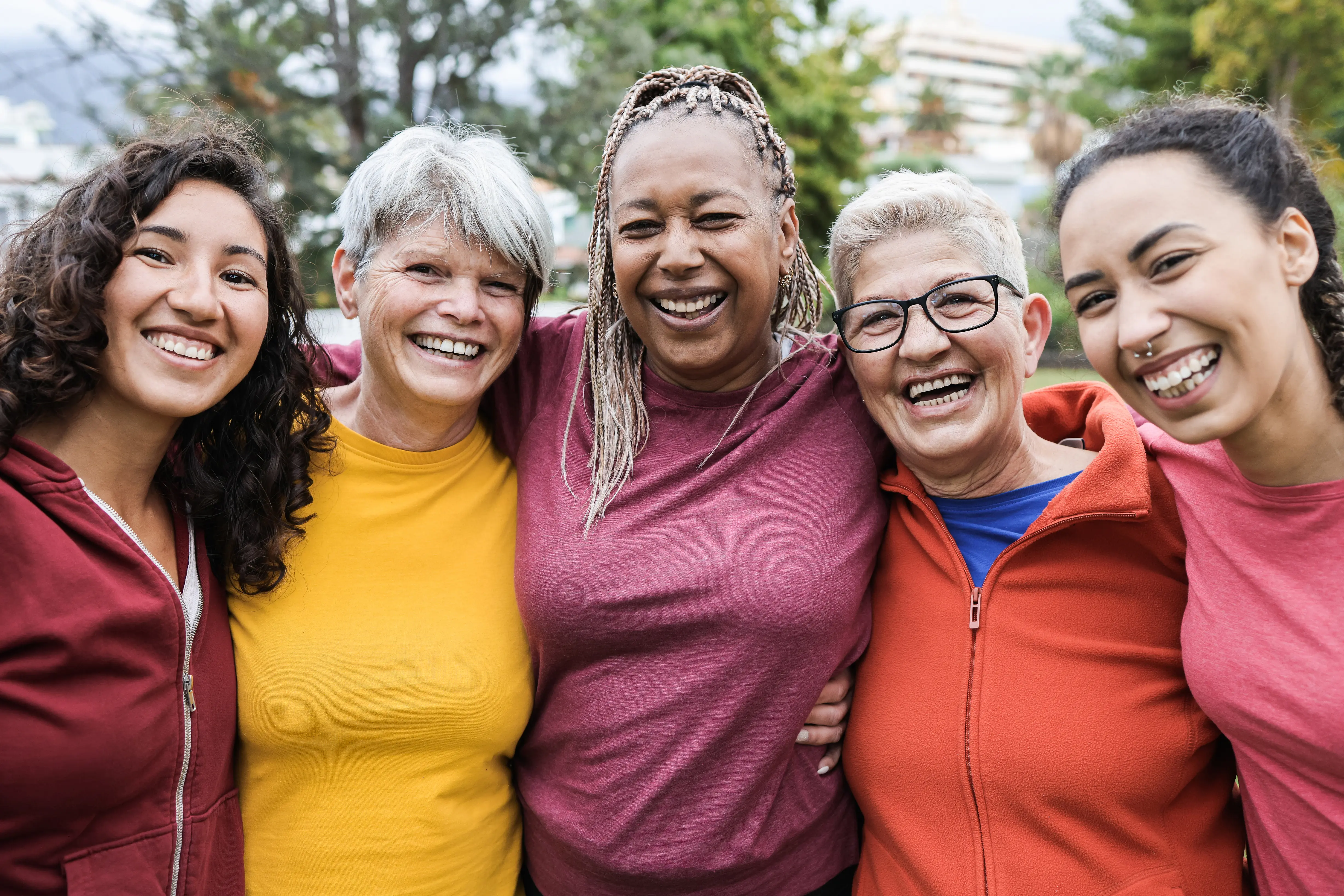 Group of five diverse women standing close together outdoors, smiling and laughing.